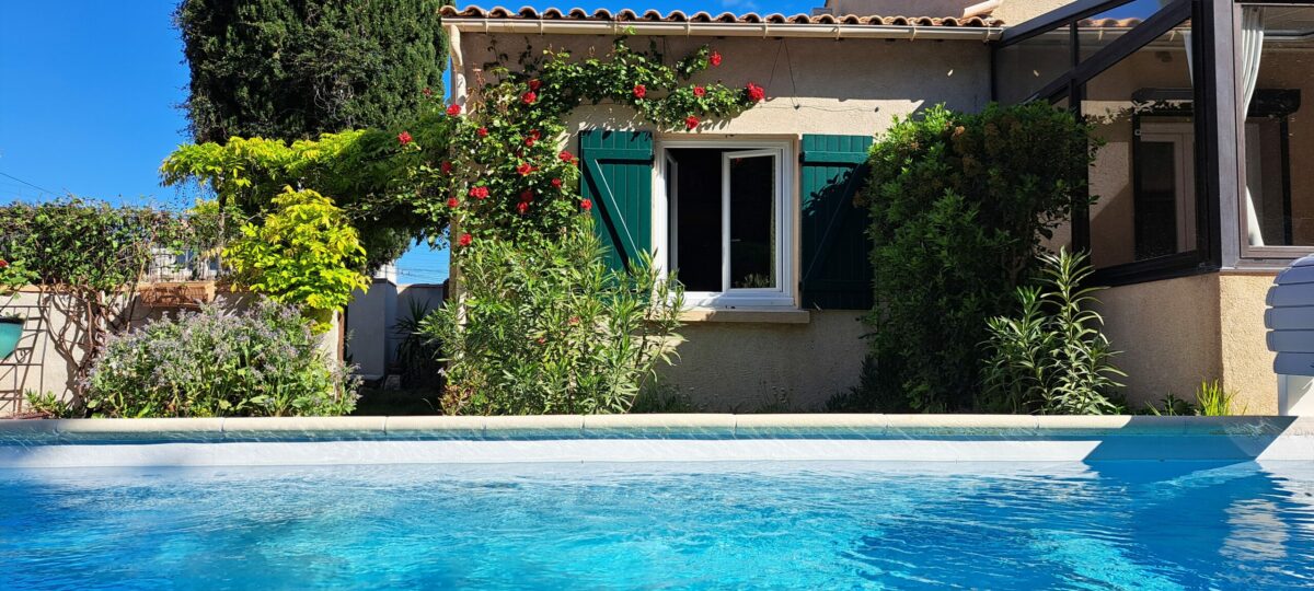 La façade sud de L’Oustau dei Aucèus et sa fenêtre aux volets verts, vues depuis le bord de la piscine partagée.
