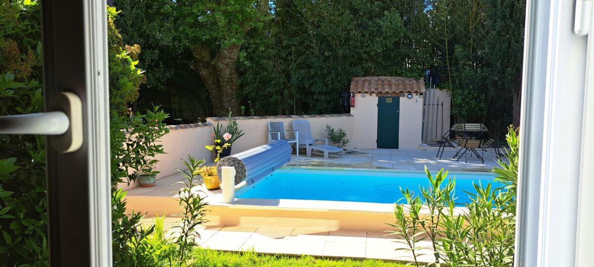 Depuis la fenêtre sud de L’Oustau dei Aucèus, votre regard s’ouvre sur la piscine extérieure partagée, son eau bleue et les arbres du jardin.