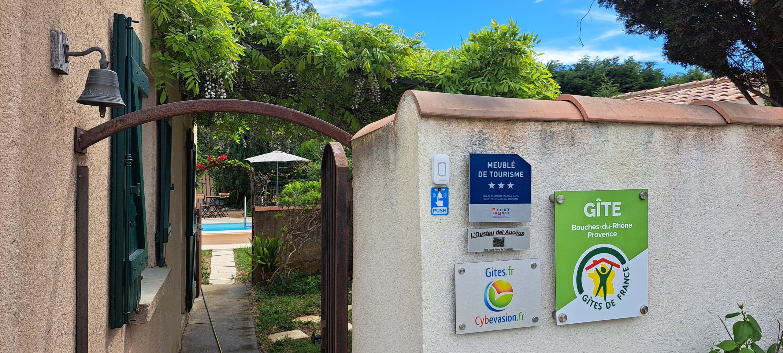 Depuis l’entrée principale de L’Oustau dei Aucèus, le regard suit la tonnelle fleurie et le passage conduisant vers la piscine extérieure partagée.