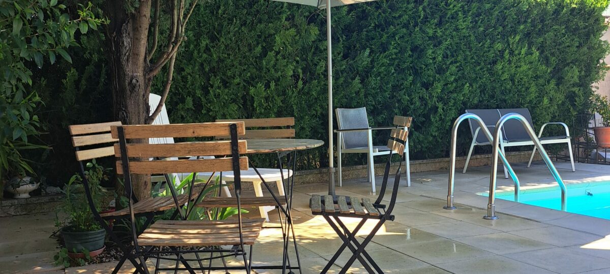 Le coin terrasse ombragé près de la piscine partagée de L’Oustau dei Aucèus, avec ses tables et ses chaises entourées de verdure.