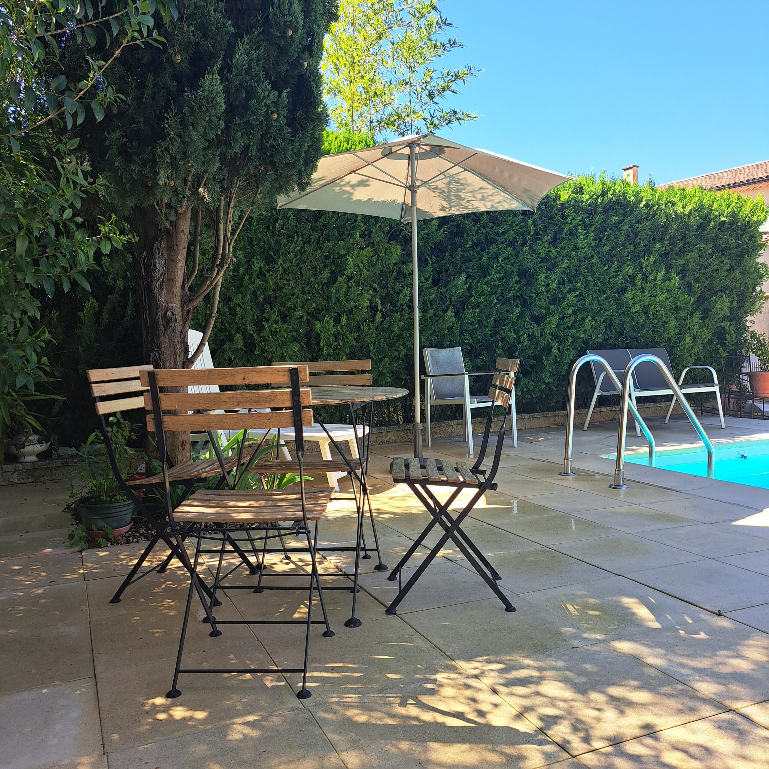 Le coin terrasse ombragé près de la piscine partagée de L’Oustau dei Aucèus, avec ses tables et ses chaises entourées de verdure.