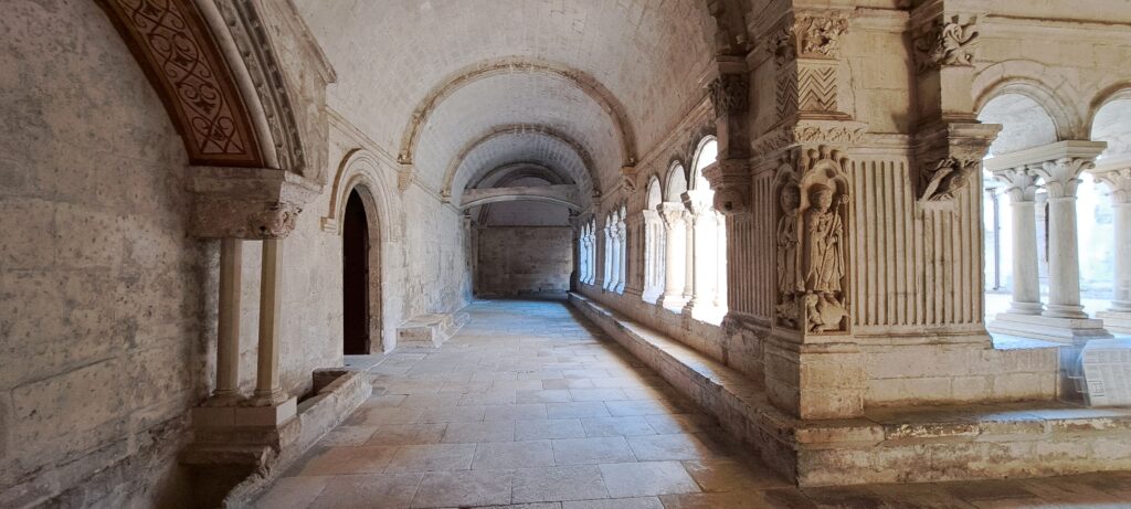 Galerie du cloître médiéval de l'Abbaye de Montmajour, avec ses voûtes en pierre, ses colonnes et ses chapiteaux sculptés représentant végétaux, figures et scènes religieuses.